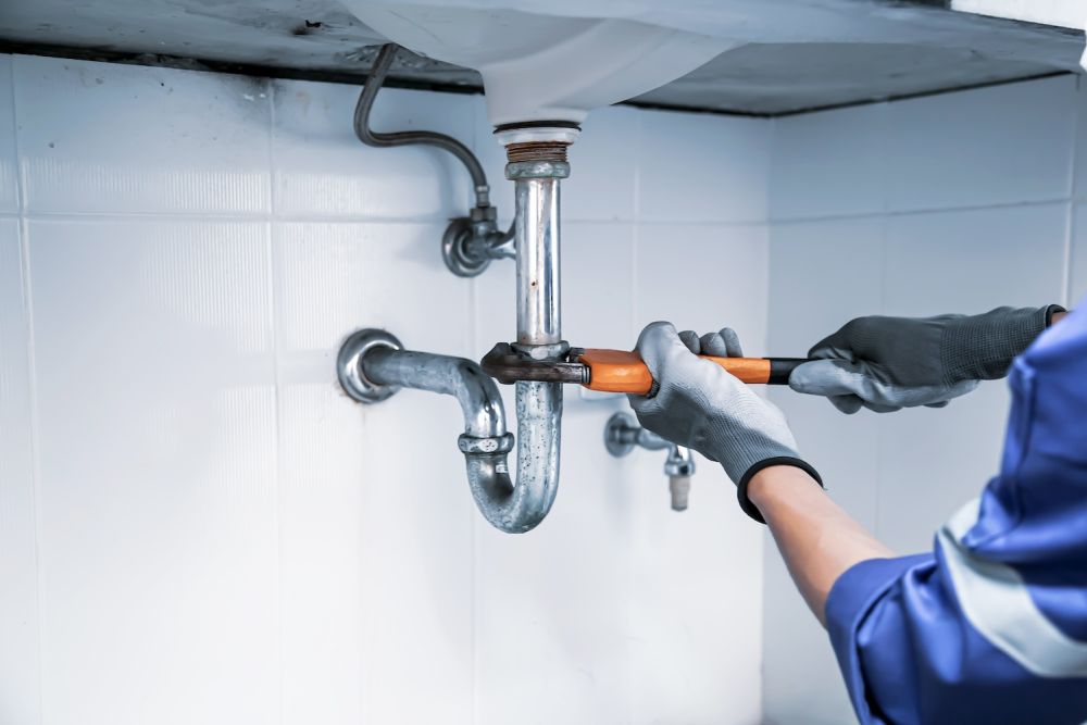 Plumber fixing under-sink pipe with wrench, wearing gloves.