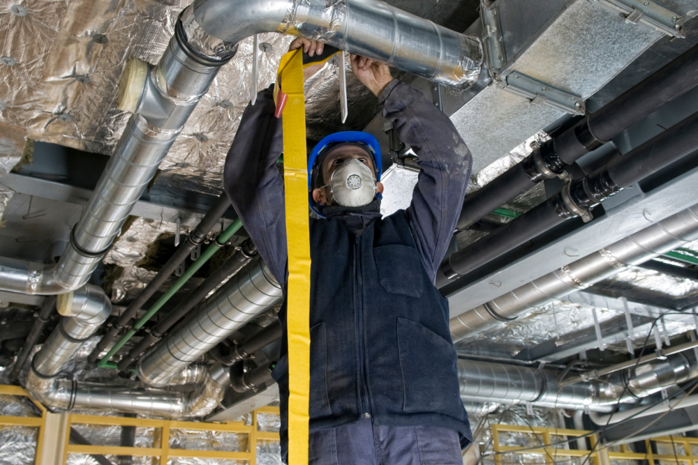 Worker in safety gear inspects HVAC ducts in industrial setting, ensuring efficient ventilation.